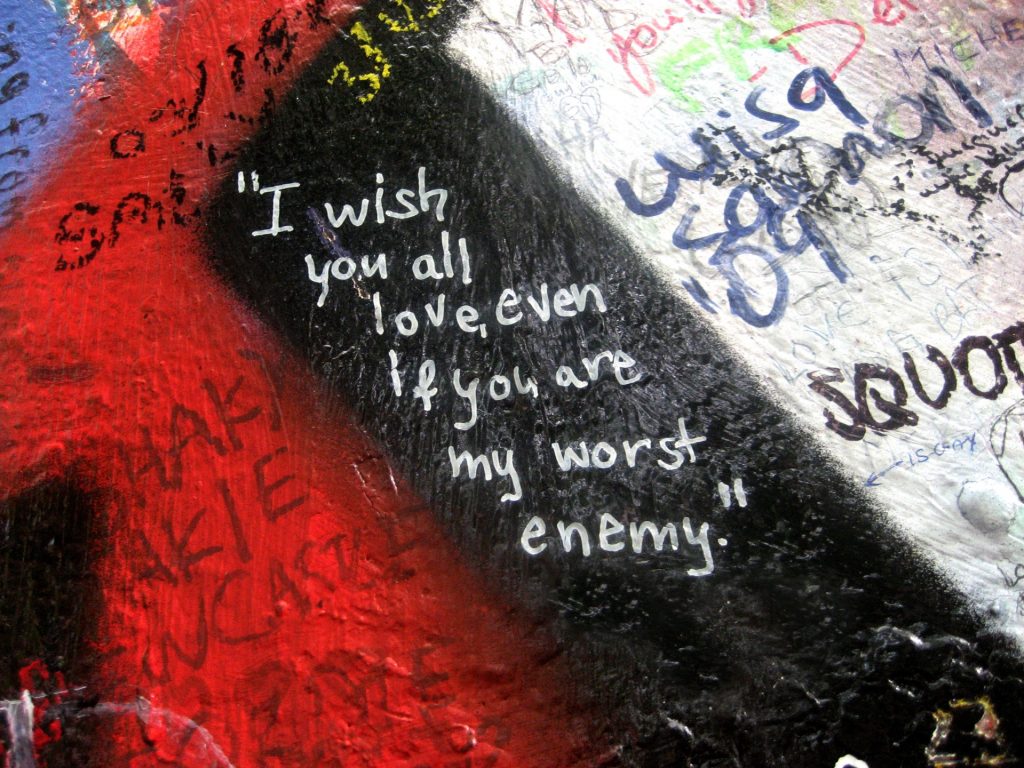 Red and black and white paint on a wall covered by various random graffiti. Center text, white on black, reads "I wish you all love, even if you are my worst enemy." 