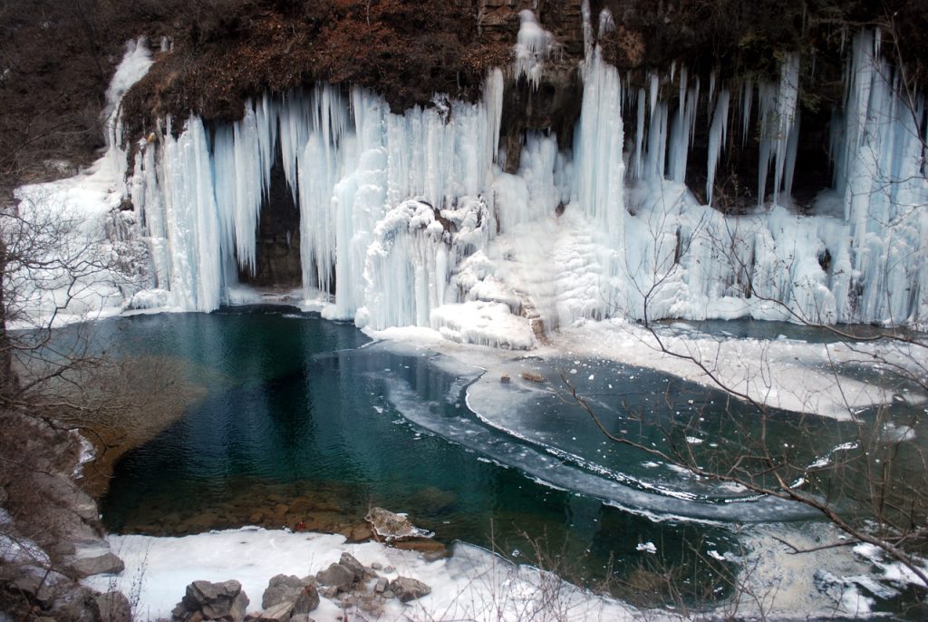 Ice falls into a rocky lake from frozen waterfalls. Barren tree branches are heavy with the weight of snow. 
