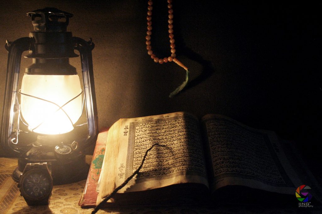 Lantern and watch on the left next to a book open to a page with Arabic script. Prayer beads hang above the book. 