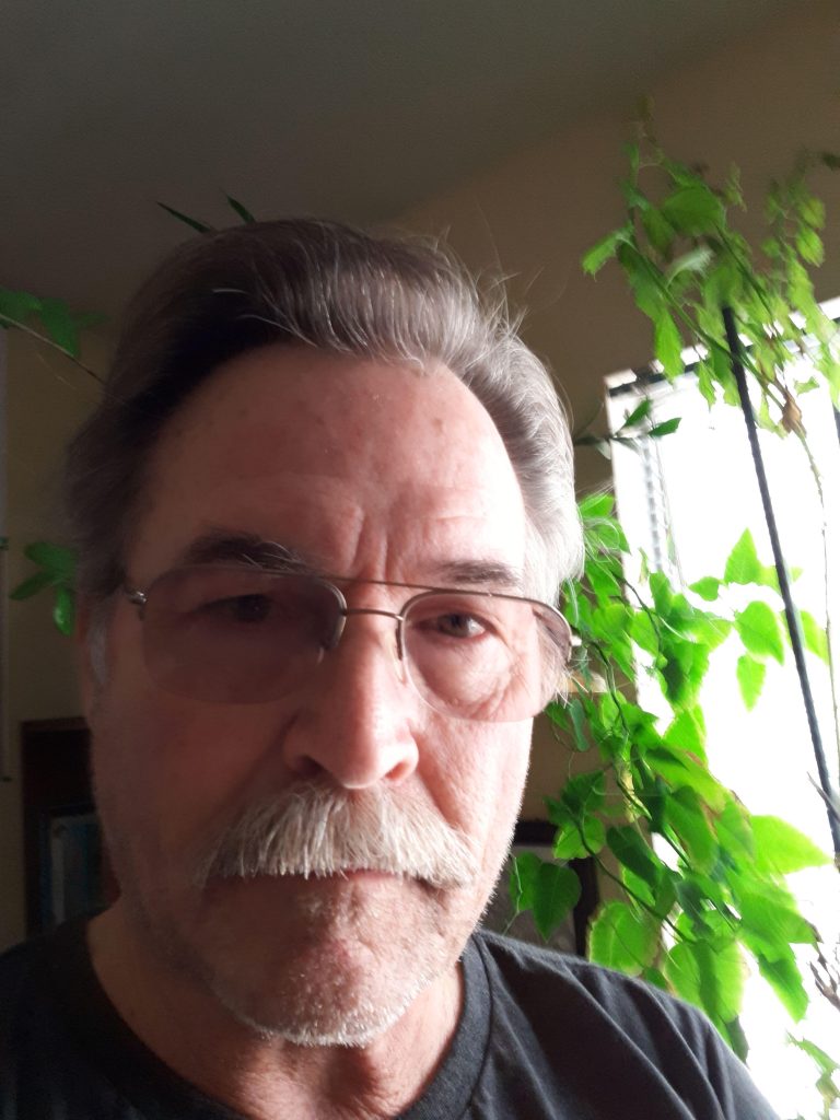 Older white man with reading glasses and a mustache and short gray hair in front of an open window and a green plant on a sunny day.