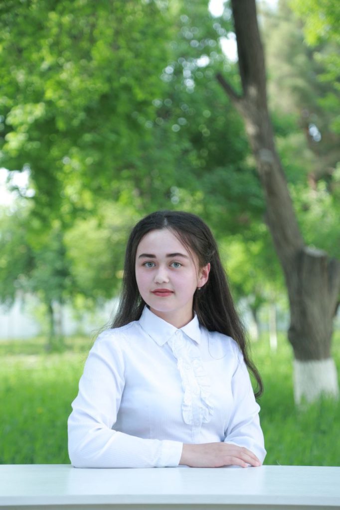 Young Central Asian woman with blue eyes and long straight brown hair and a white collared shirt with ruffles down the middle posing in front of a blurry background of grass and trees. 