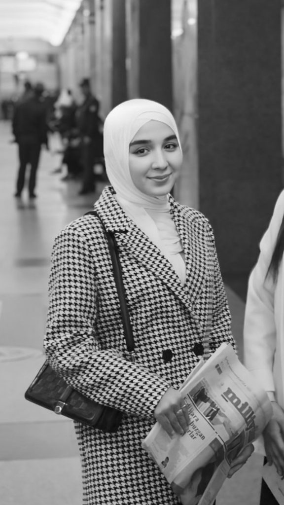 Black and white photo of a Central Asian young woman with a white headscarf and black and white patterned sweater. She's in a hallway with other people in the background and is holding a newspaper. 