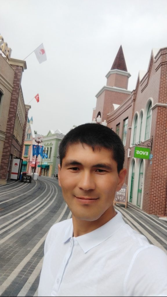 Young Central Asian man with short brown hair, brown eyes, and a white collared shirt in the street with concrete pathways and brick buildings on either side. 