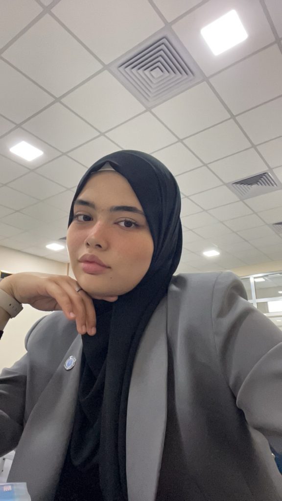 Central Asian teen girl with a black headscarf, brown eyes, a gray coat, and a wristwatch. 