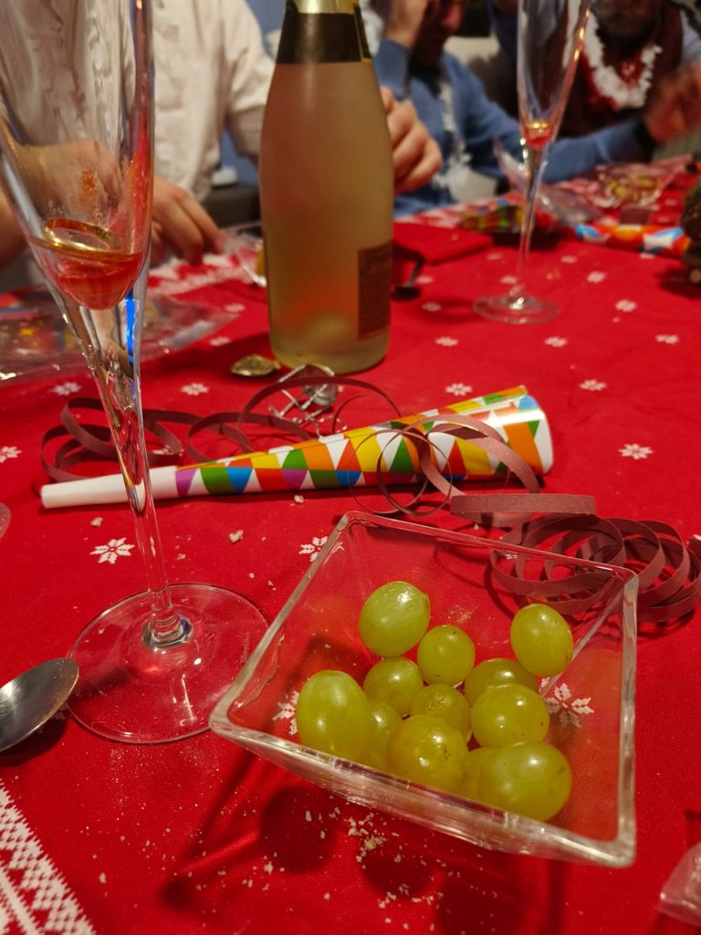 Twelve green grapes in a glass bowl on a red tablecloth with a party noisemaker and a bottle of champagne and two glasses. 