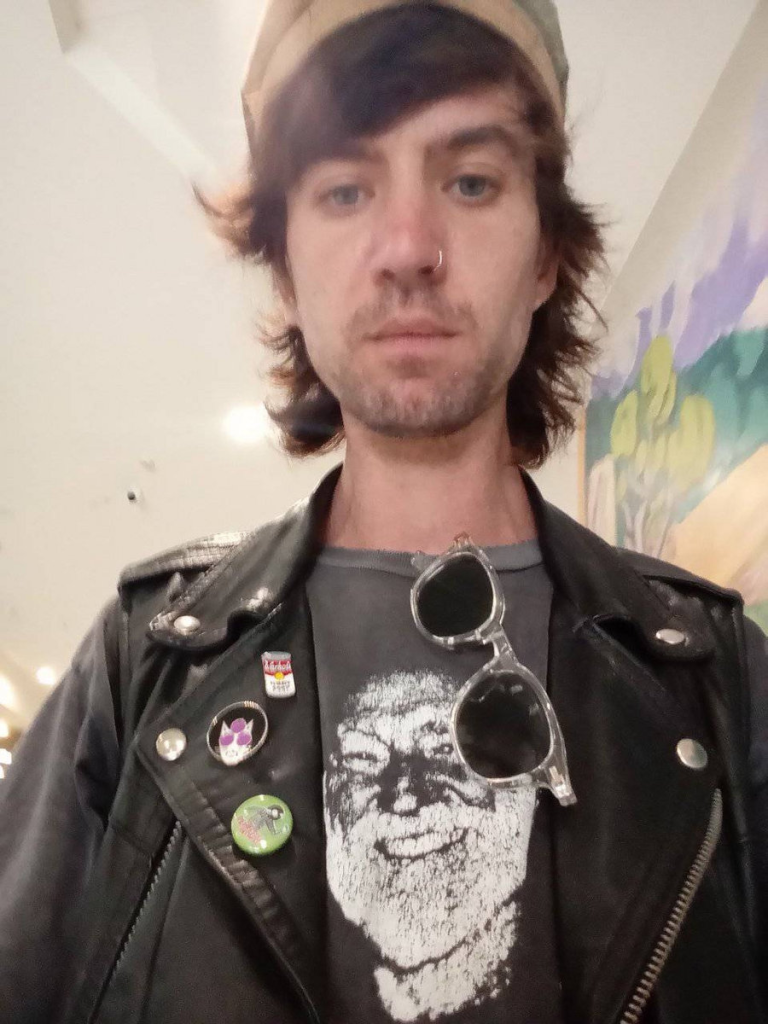 Young (20-something) white man with a nose ring, short scruffy hair, black leather jacket with pins on the lapel, sunglasses, and a gray tee shirt with the face of an old man on the front.