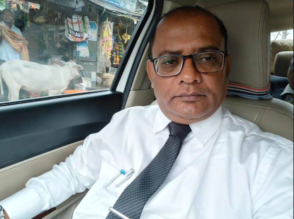Older middle aged South Asian man with reading glasses and a white collared shirt and black spotted tie in a car with a street market and white cow out the window.