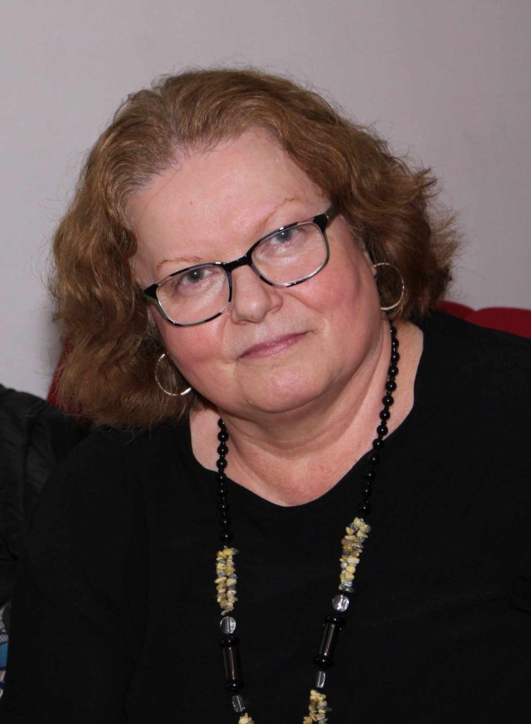 Light skinned Eastern European older middle aged woman, with reading glasses, hoop earrings, and brown curly shoulder length hair. She's got a black top and a black and brown necklace. 