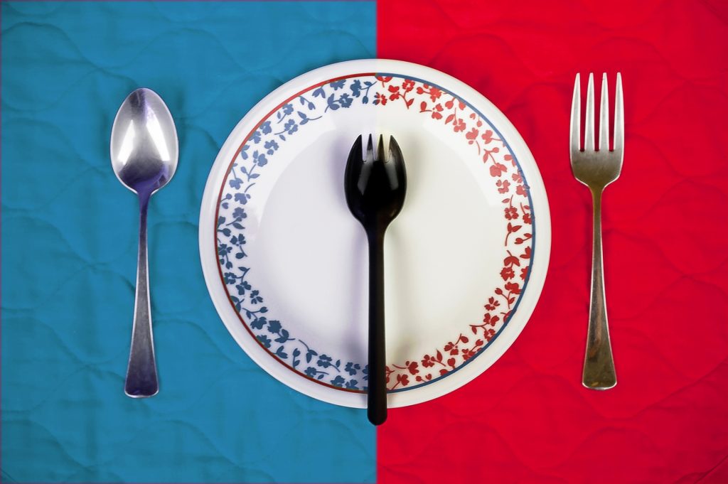 Image of a spoon on the left of a plate with a blue design and blue tablecloth and a fork on the other side next to the other half of the plate with a red design. A black plastic spork with tines at the end of a spoon is in the middle of the plate. 