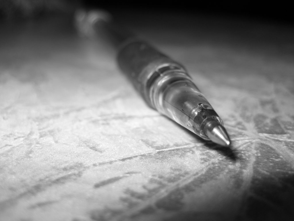 Ballpoint pen at an angle photographed up close, pointing to the viewer. On a concrete table. Photo is black and white. 
