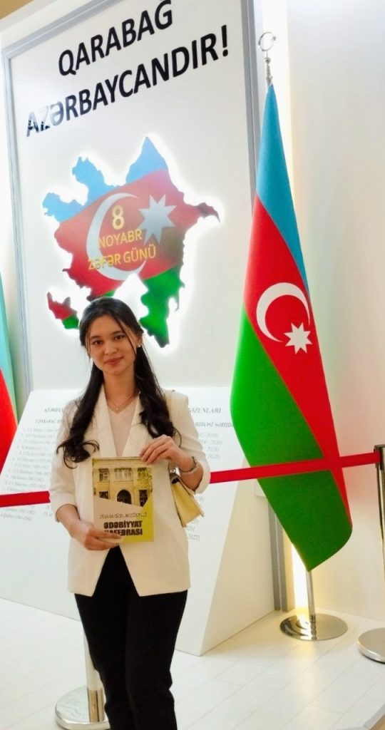 Young Central Asian woman with long straight dark hair and a white coat, blouse, and black pants holds a book and stands in front of a patriotic Azerbaijani mural and flag. 