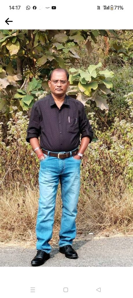 Older middle aged South Asian man with reading glasses, a dark collared shirt, and blue jeans. He's outside in front of a tree and dry grass. 