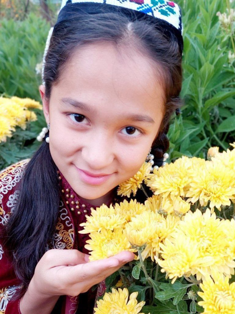 Young teen Central Asian girl with a colorful embroidered headdress, brown eyes, a traditional garment with orange and white stitching on deep burgundy cloth, holding yellow flowers outside. 