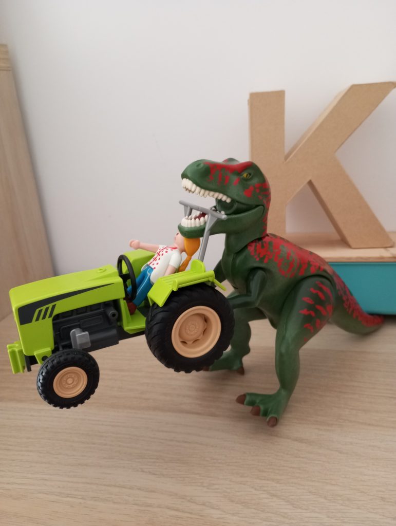 Green and red plastic dinosaur holds a light green tractor with a plastic female driver figurine. 