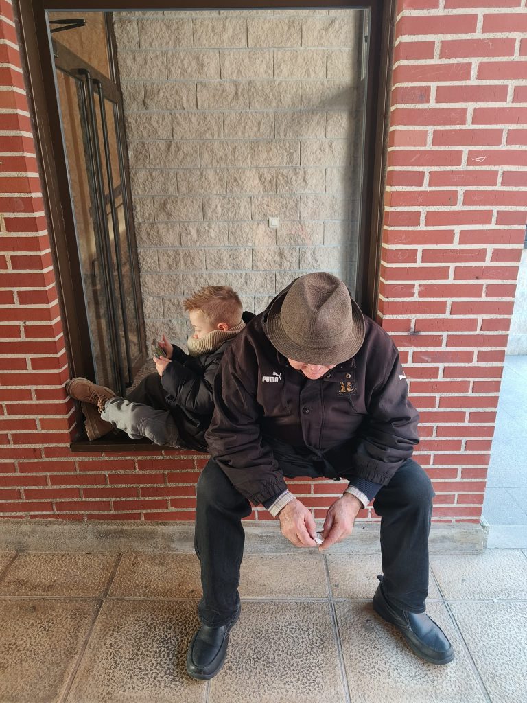 Color photo of an older white man with a brown hat, black coat, black pants and dress shoes sitting on a windowsill in a brick building. A boy is next to him, facing to the side, looking at a rock or phone, something in his hands. 