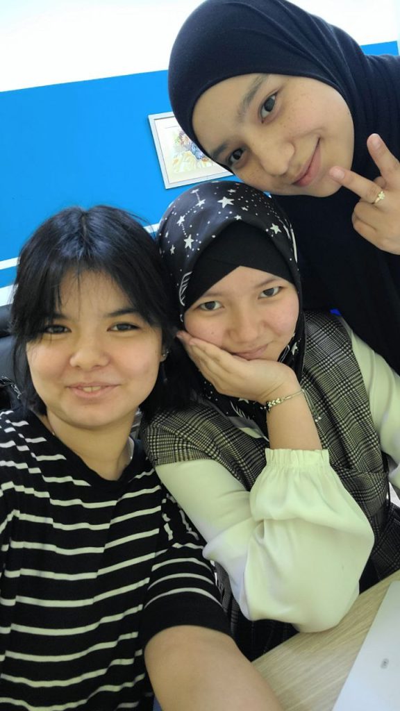 Three Central Asian young women's heads in the photo, two with headscarves and the other without. They're in a shawl, a blouse and best, and a striped shirt. 