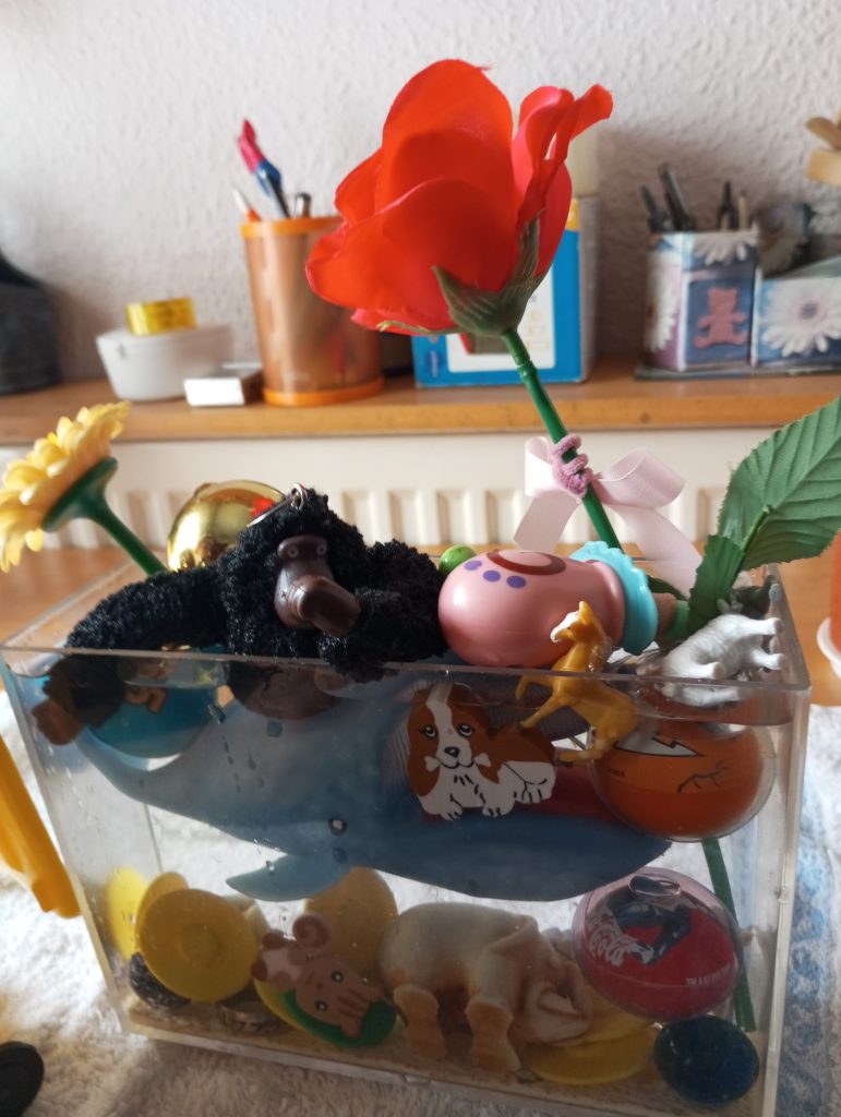Plastic red rose in an aquarium filled with a plastic whale and various kids' toys. Shelf in background with jars of pens. 