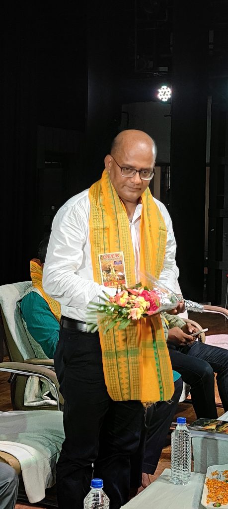 Older South Asian gentleman with a yellow scarf, white collared shirt, and black pants. He's standing up near a chair holding a bouquet of flowers and his book. 