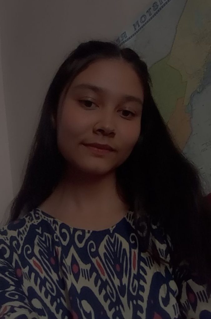 Central Asian teen girl with long dark hair in a floral blue and white blouse standing in front of a map.
