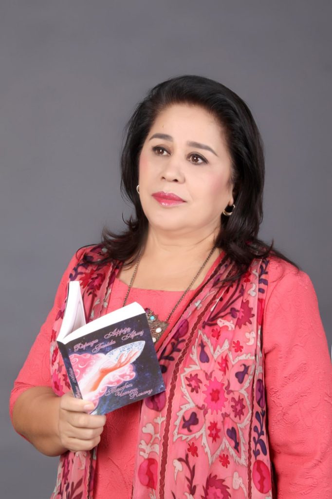 Middle aged light skinned Central Asian woman with shoulder length hair and earrings wearing a pink floral blouse and holding a book. 