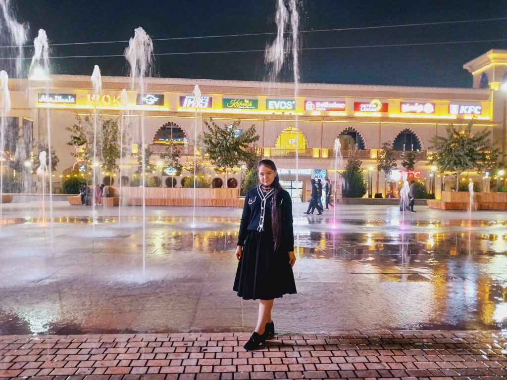 Teen Central Asian girl in a coat with buttons, a scarf, long dark hair, a skirt, and boots. She's outside at night in front of a concrete fountain and a variety of restaurants. 