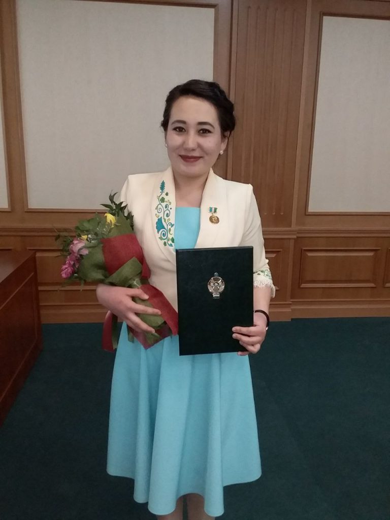 Middle aged Central Asian woman with dark hair up in a bun, an off-white coat with a medal and a floral design, and a light blue dress. She's holding an award and a bouquet of flowers. 