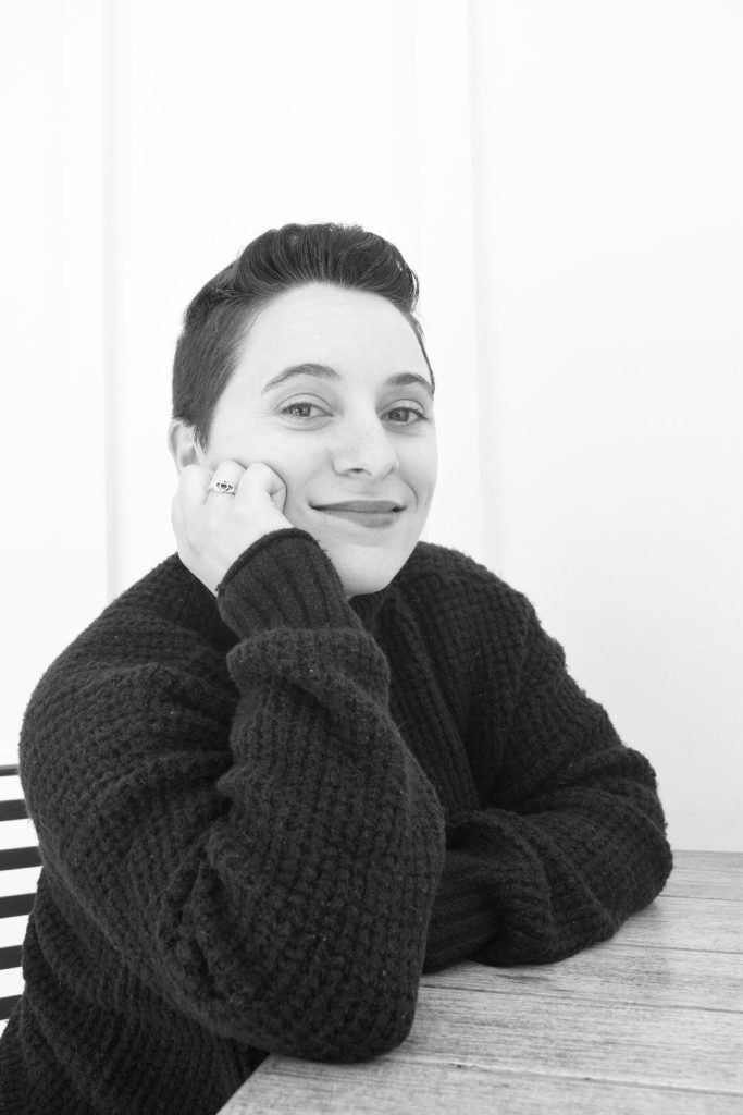 Young seated white woman with short hair, brown eyes, her elbow on the table and her head resting on her hand. She's got a dark colored sweater and a ring on her finger. 