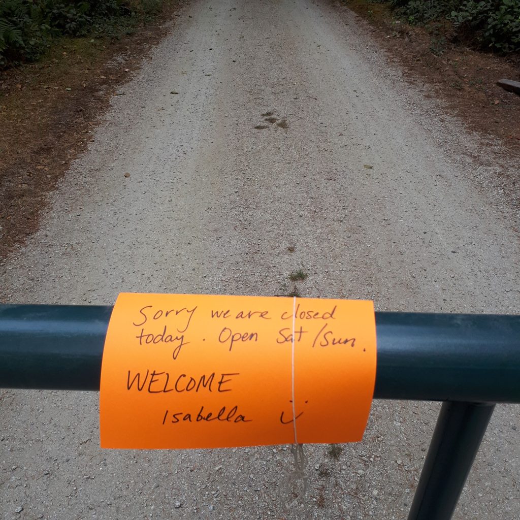 Yellow sticky note saying that a museum is closed, from Isabella, on a metal gate over a gravel trail. 