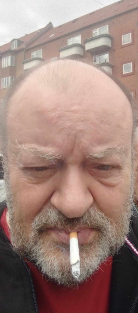 Old white guy with thinning hair and a beard in a red shirt and black coat smoking a cigarette. He's in front of a brick building with windows and balconies.