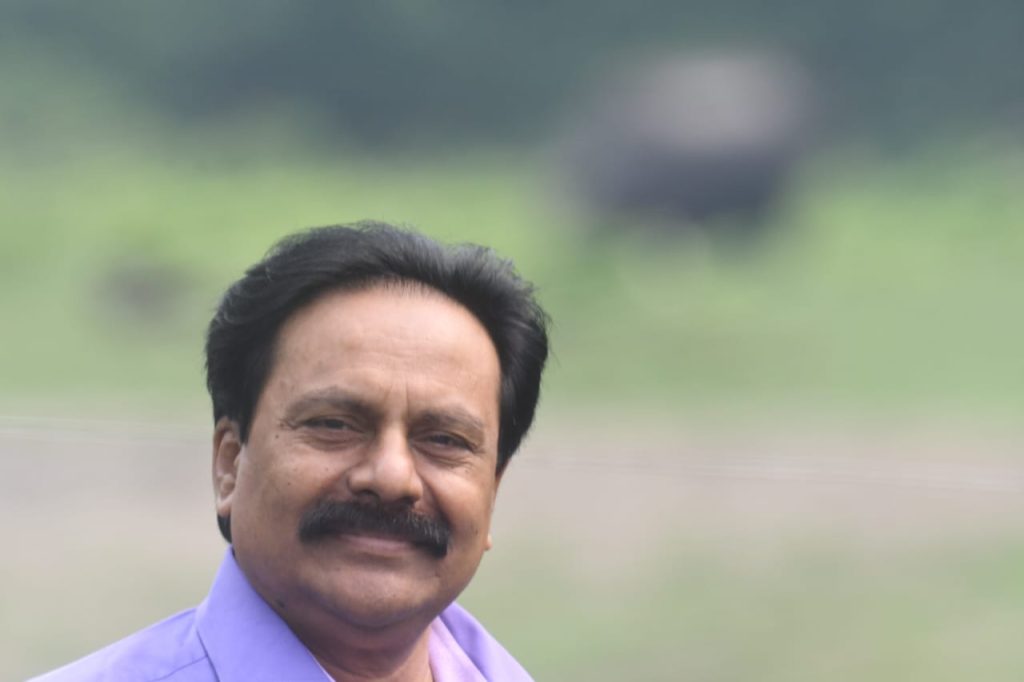South Asian man with short brown hair, a brown mustache, and a purple dress shirt in front of a hazy green outdoor background. 