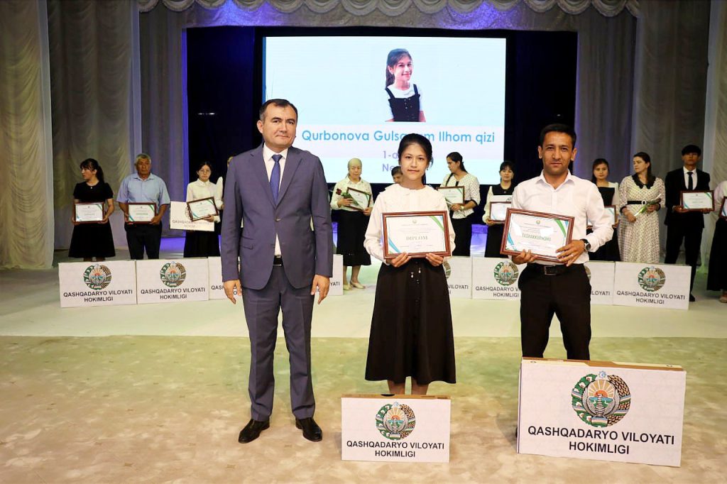 Two college-age Central Asian students, a guy and a girl, in white collared tops and a skirt and slacks, standing next to an older man in a gray suit and blue tie. The students hold framed certificates as do others behind him. Our author, Gulsanam Qurbonova's, face is up on a screen. 