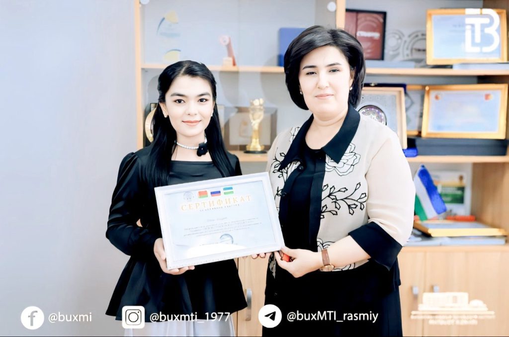 Young teen Central Asian girl with long dark hair and a white necklace and black top and white skirt holding a certificate. An older middle aged woman, likely a teacher, in a black dress and light colored jacket, presents it to her. They're in a classroom with awards in a wooden case behind them. 