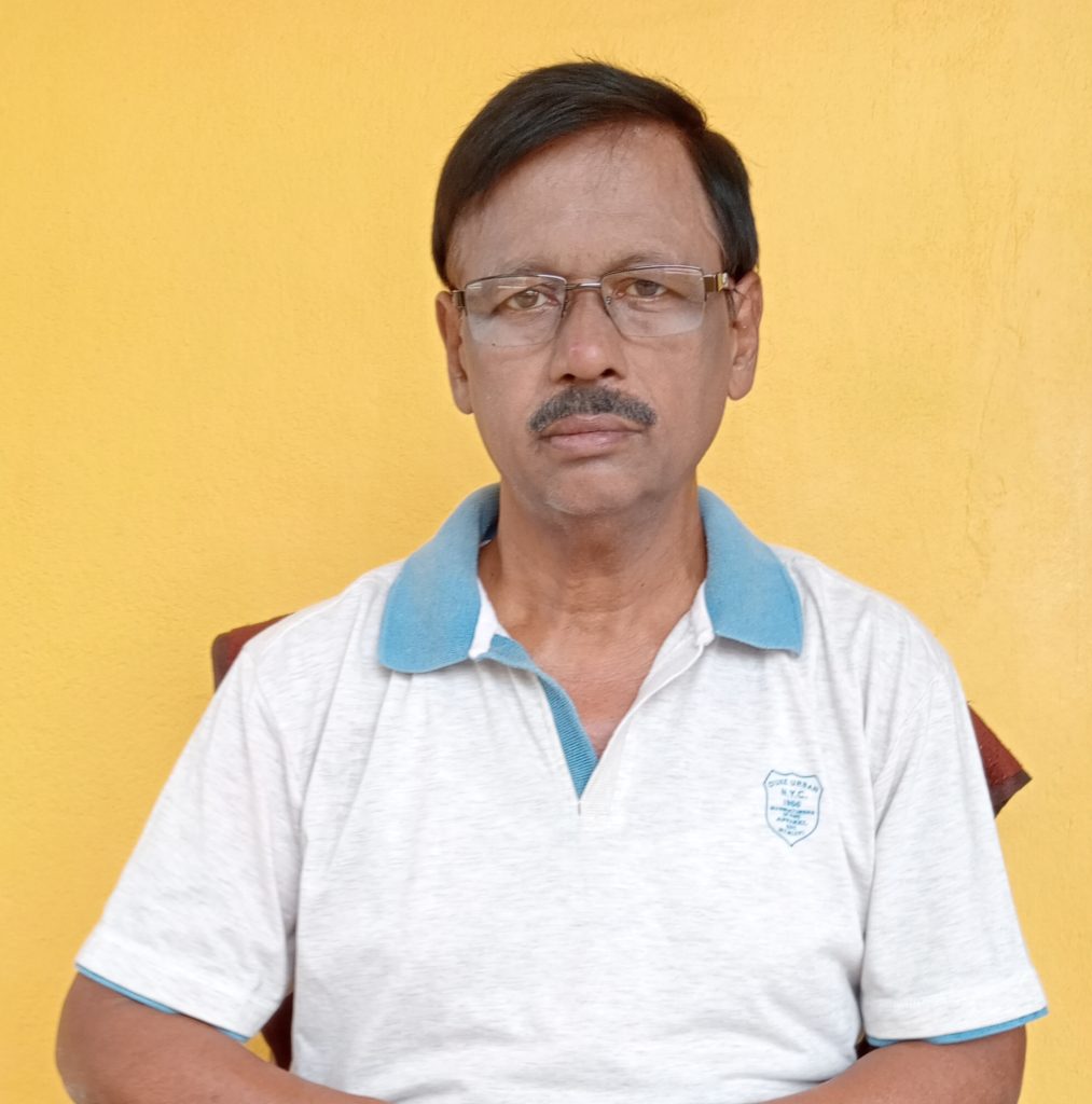 Older middle aged South Asian man with reading glasses, short brown hair, a white collared shirt with blue collar. He's sitting in front of a yellow background. 