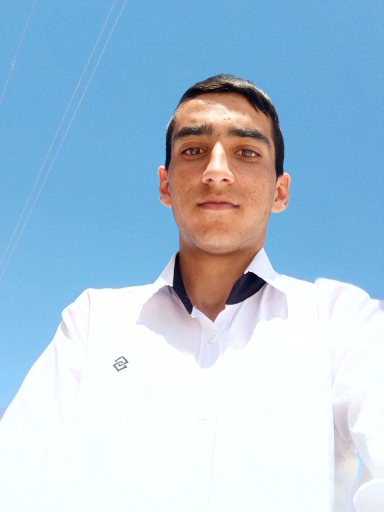 Young Central Asian man with short brown hair, brown eyes, and a white collared shirt out in a sunny day in front of blue sky. 