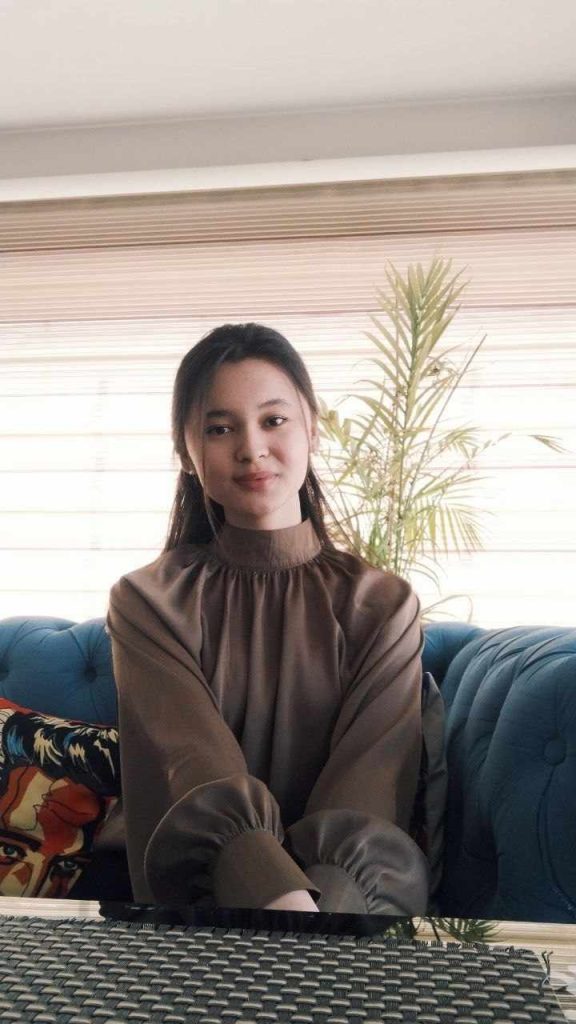 Central Asian teen girl with a brown ruffled blouse and long dark hair in front of a fern houseplant and a window with blinds. 