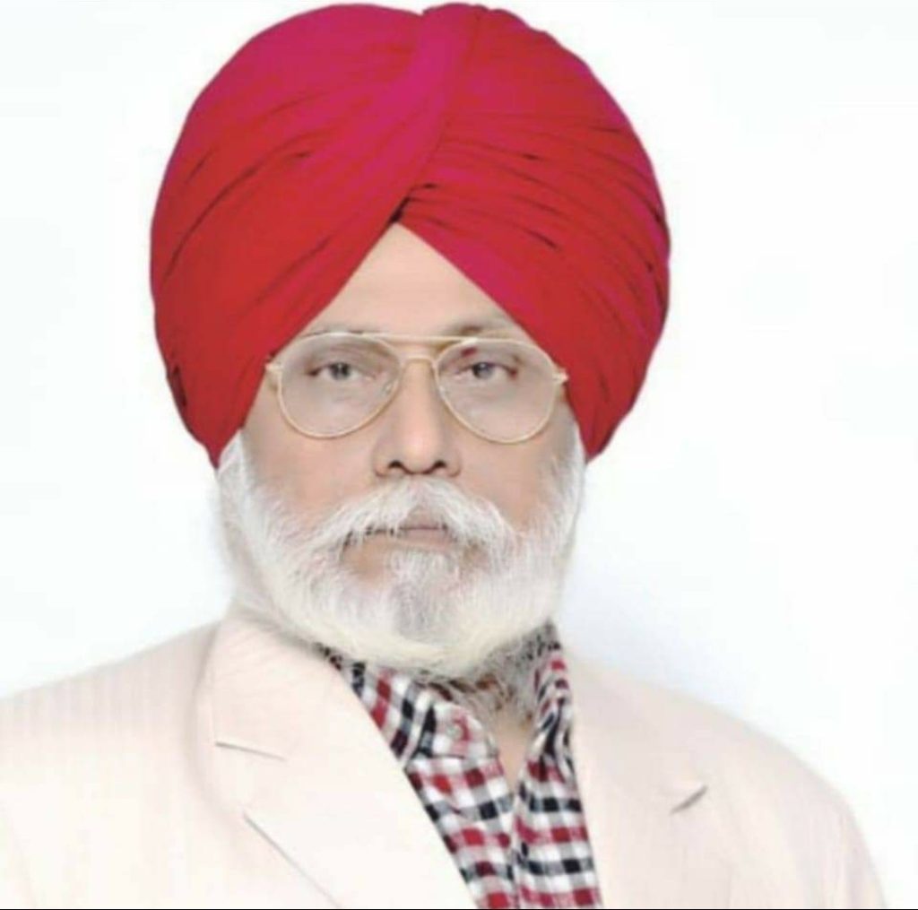 South Asian man with a red turban, reading glasses, and a white beard, plaid collared shirt, and gray coat. 