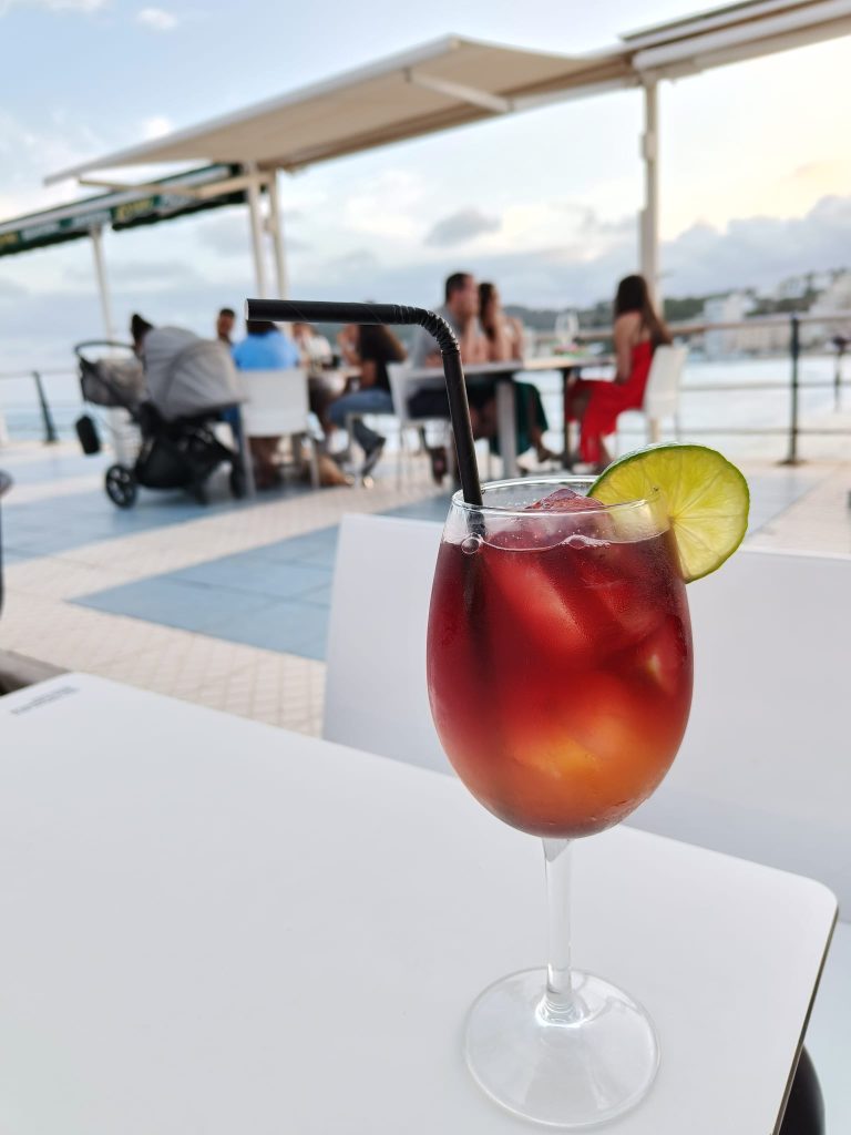 Red drink in a glass with ice and a lime slice and a black straw on a white table in an outdoor cafe. People, including a woman in a red dress and a family with a baby stroller, sit in the distance. Some clouds in the sky. 