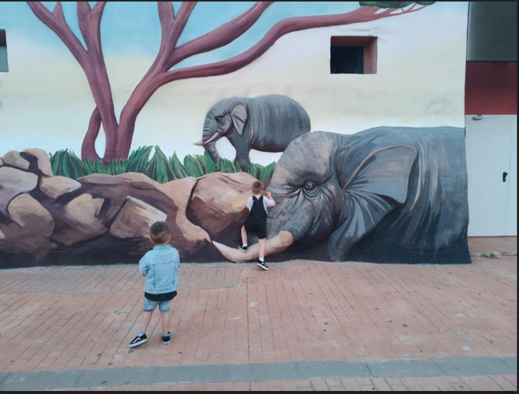 Little boy, light skinned with tennis shoes, short brown hair, and a blue shirt and shorts, standing in front of a mural of two huge elephants and rocks and grass and a tree. Another boy, who looks similar, is up front touching the wall. 