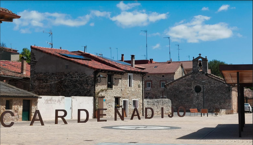 Stone buildings with red tile roofs in a stone city square. Sunny day with a few clouds in the sky, some chairs in the distance. 