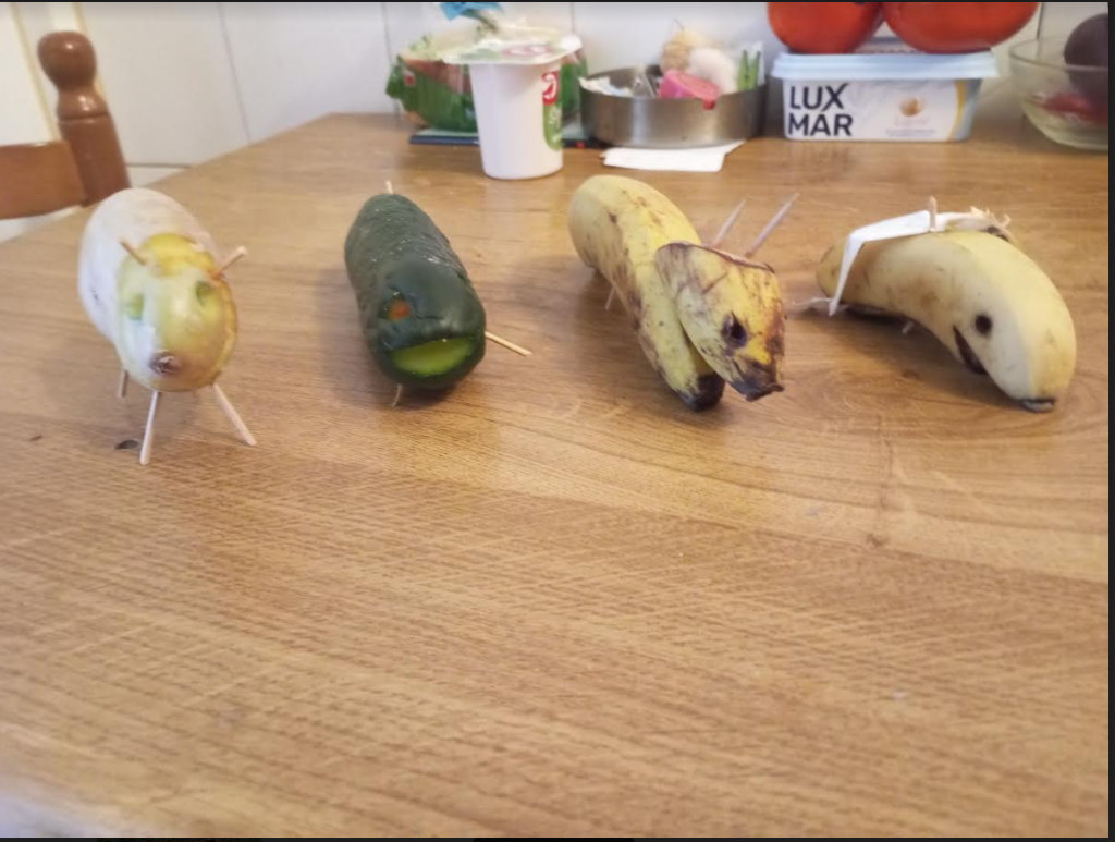 Animal figures made of bananas, a cucumber, and a potato, propped up with toothpicks. They're on a wood table with some other kitchen items in the distance. 