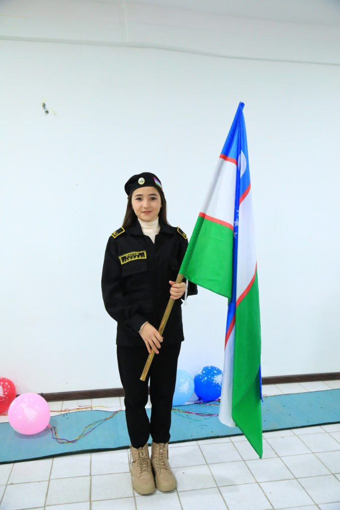 Young Central Asian woman with a hat and black coat and black pants and white collared shirt underneath holding a yellow, white and blue flag.