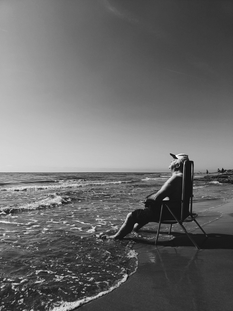 Black and white photo of an older man sitting in swim shorts on a beach chair facing into the ocean. 