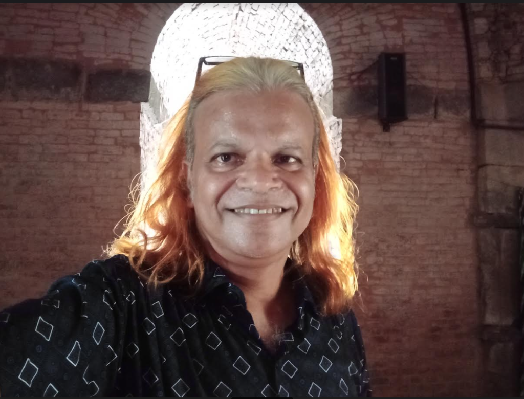 South Asian man with shoulder length white and red-orange hair, a black and white top, brown eyes, and reading glasses at the top of his head. He's standing in front of an exit to a brick vault building with light behind him. 