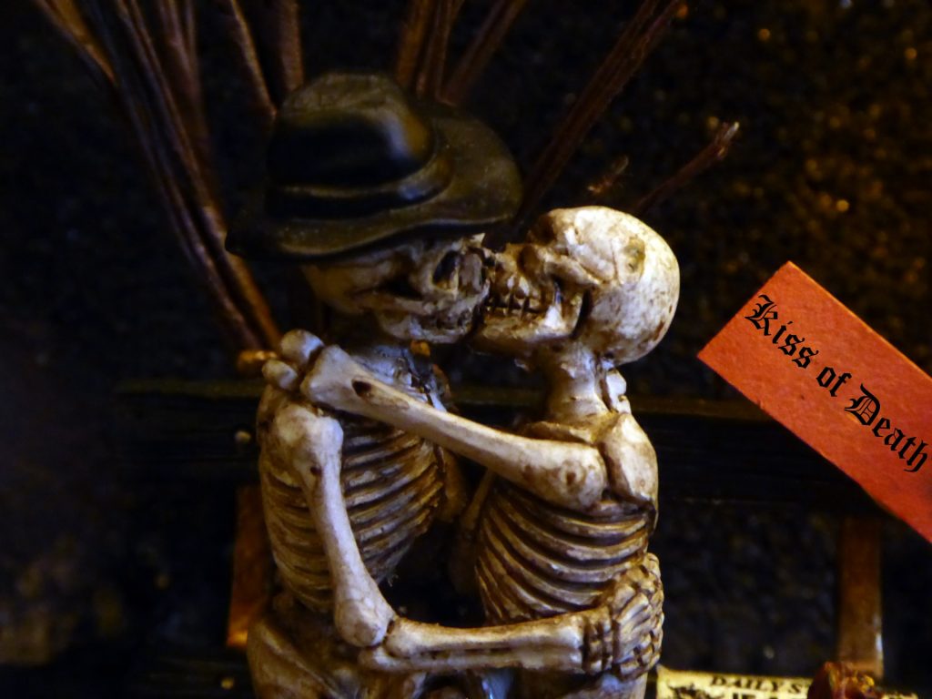 Two skeletons dancing, one with a top hat, with a sign in the background saying Kiss of Death.