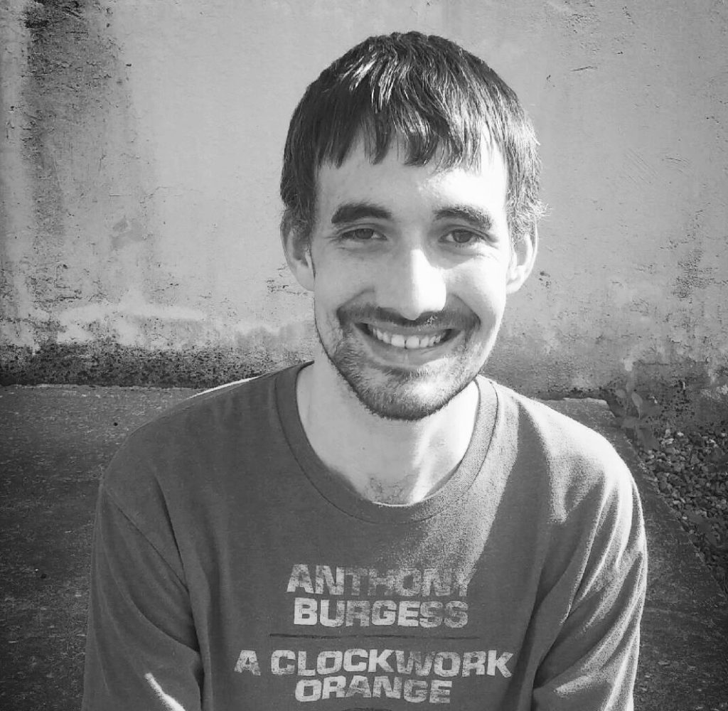 Young skinny white guy (in his 20s) with short brown hair, a small beard and mustache, and a dark colored tee shirt reading "Anthony Burgess' A Clockwork Orange." Black and white photo, he's on a patio in front of a concrete wall. 