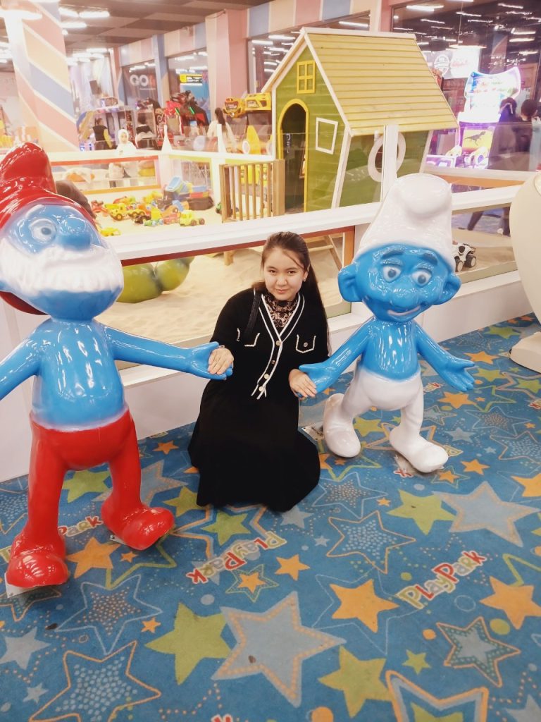 Blue lifesize Smurf characters and a toy gingerbread house surround a young teen Central Asian girl with a black coat with white trim and long dark hair. 
