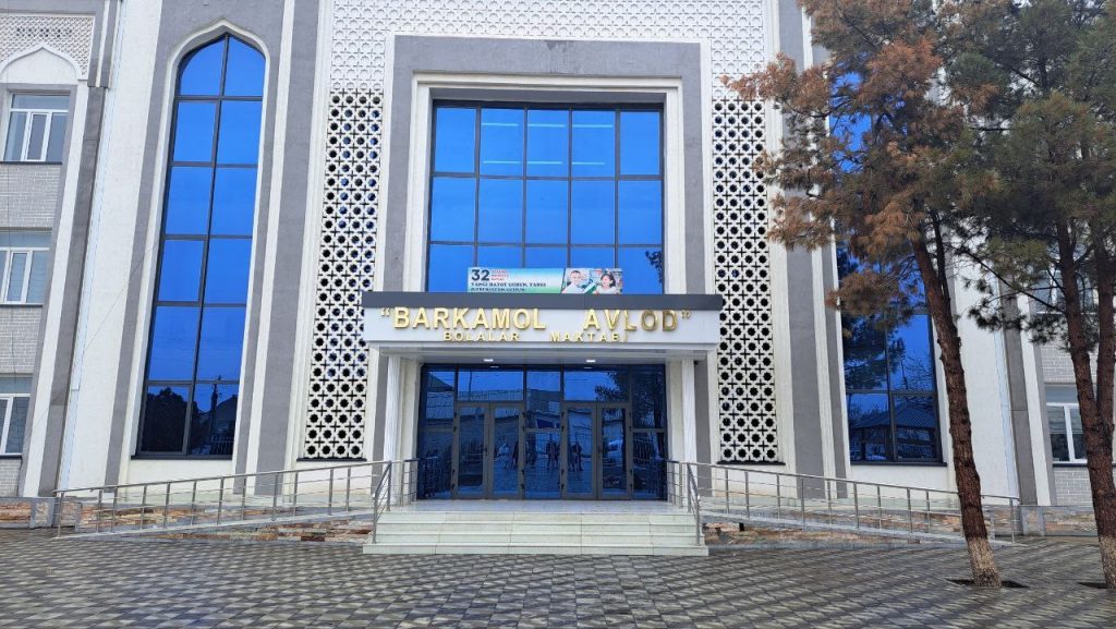 Barkamol Avlod Children's School. Large urban looking building with large glass windows and big gold letters. Some trees and a concrete walkway in front. 