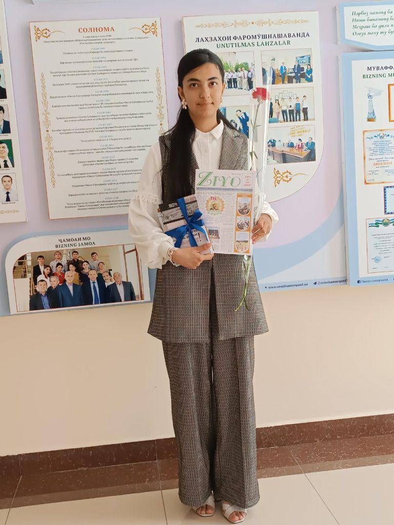 Central Asian teen girl with long dark hair, brown eyes, and a black and white checkered school uniform vest and pants over a white collared shirt. She's holding books and a rose and standing in front of posters on the wall in a schoolroom. 