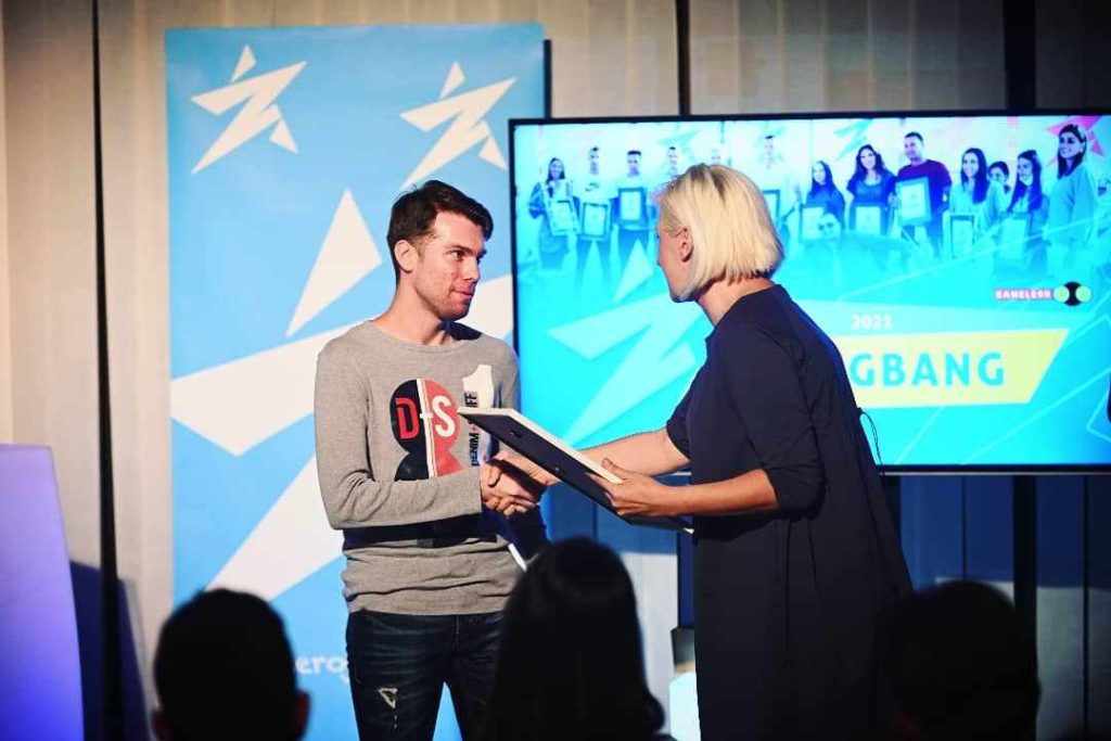 Young white guy in a gray sweater and dark pants with short brown hair up on stage receiving an award. 