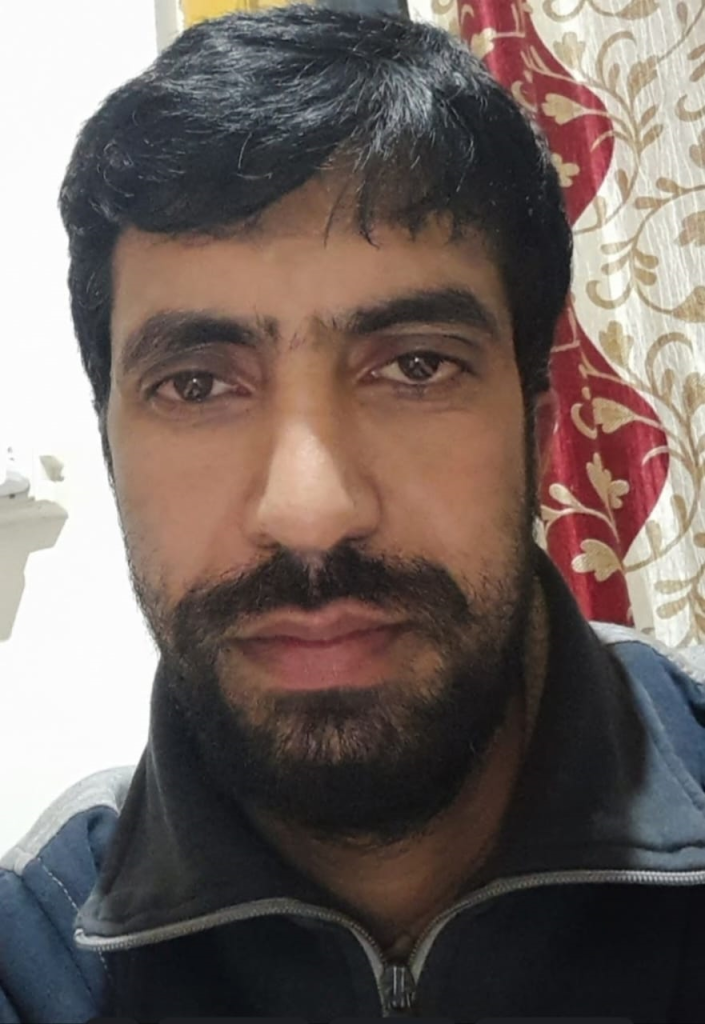 Young South Asian man with short brown hair and a dark trimmed beard and mustache. He's got a blue coat with a zipper. Behind him is a red and white and tan floral curtain. 